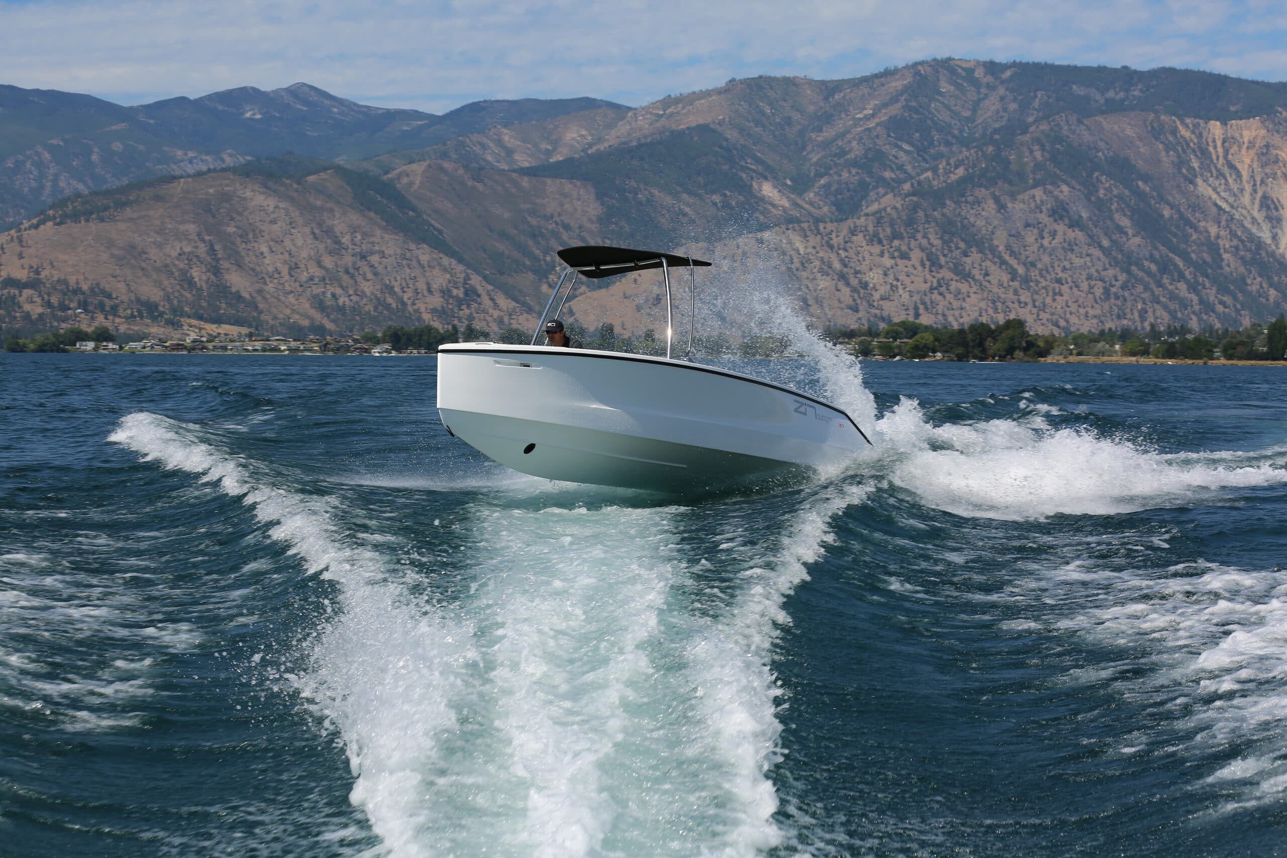 A white boat (2026 Zin Boats Z7) is planning through a wake in the open water. There are mountains in the background.