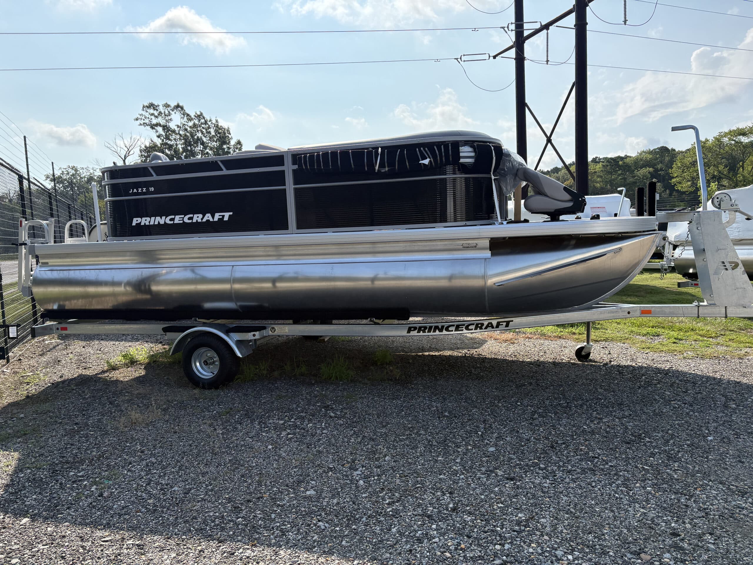 Side view of a Princecraft (2026 Princecraft Jazz 19 4s) pontoon boat on a trailer, showing the port side, railing, seating, and bow.